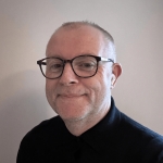 Chris O'Donnell - Chris has short hair, wears black glasses and a black top.