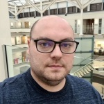 Matt Barnard - Matt is standing in an atrium. He's wearing black glasses and a dark-coloured top