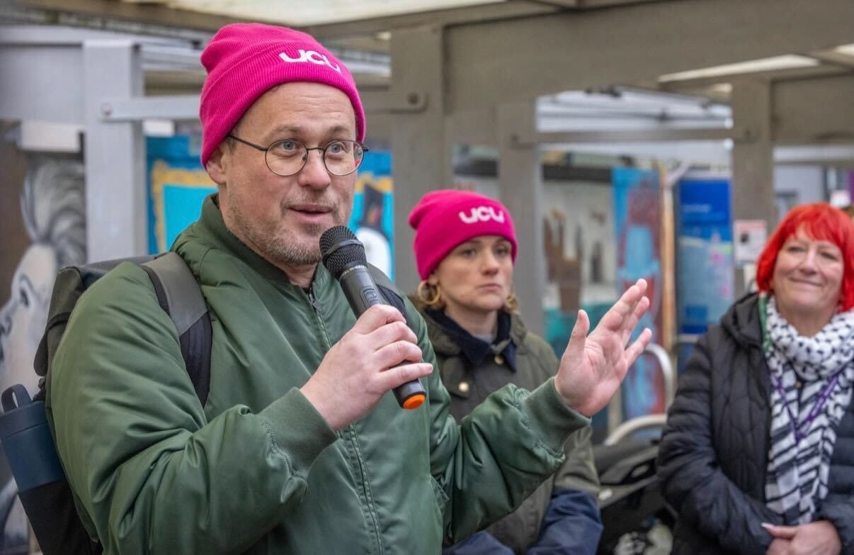 A white man in a pink UCU beanie speaks into a microphone. Two women are in the background.