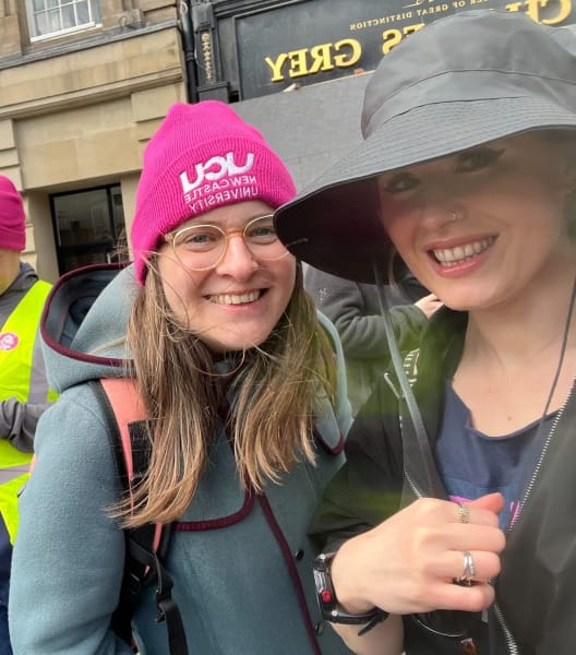 Two women, one in a pink UCU beanie, stand under an umbrella smiling at the camera