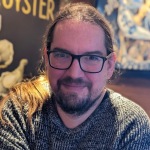 Rob Clarke. Rob has long brown hair  tied back in a pony tail, a beard and wears dark-rimmed glasses. He's wearing a woolly jumper.
